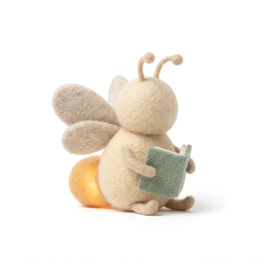 A felt firefly in cream wool with a glowing amber tail, reading a small sage-green notebook